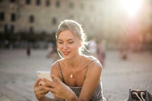 Smiling woman using smartphone outdoors in city square with sunlit backdrop.